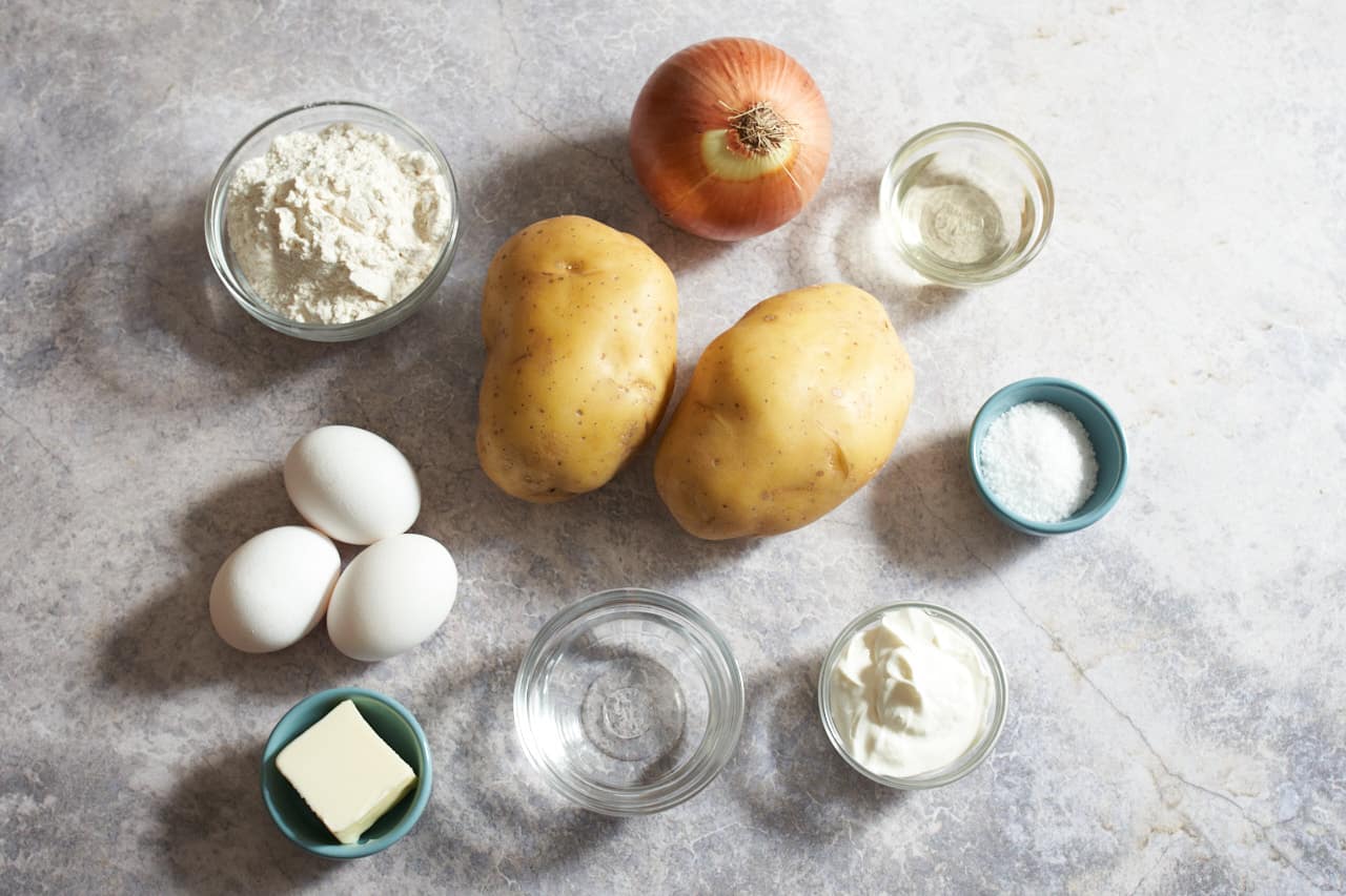 Overhead shot of various ingredients on a grey countertop, including flour, potatoes, eggs, an onion, butter, salt, oil, water, and sour cream.