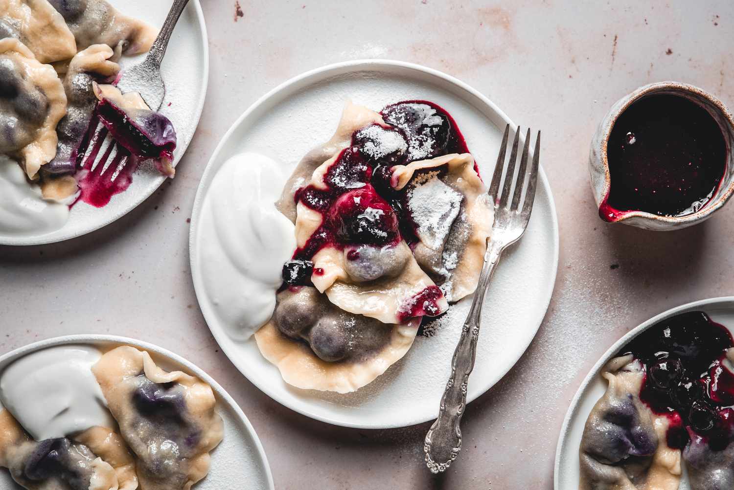 Four white plates with blueberry varenyky, served with a dollop of sour cream and a dusting of powdered sugar. A silver fork rests on one plate, and a small pitcher of blueberry sauce is placed on the side.