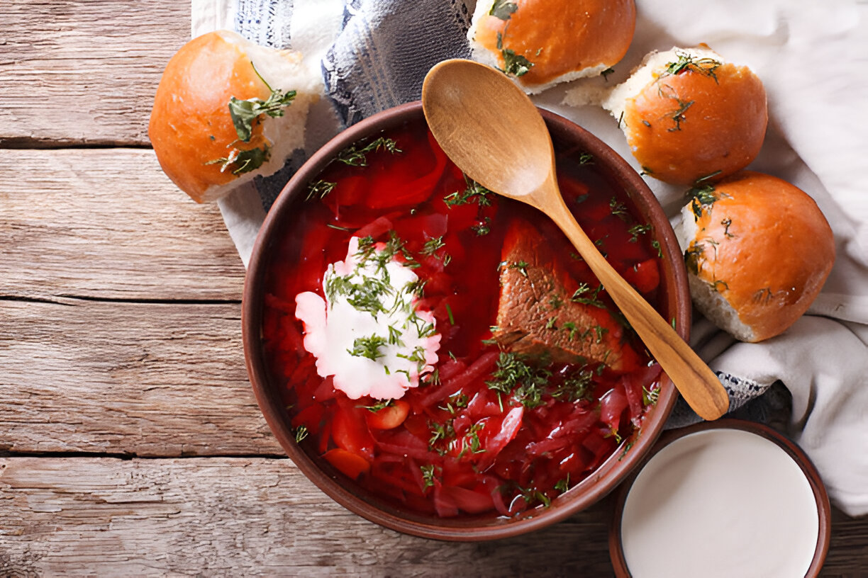 A bowl of borscht garnished with fresh dill and sour cream, served with small buns on a wooden table.