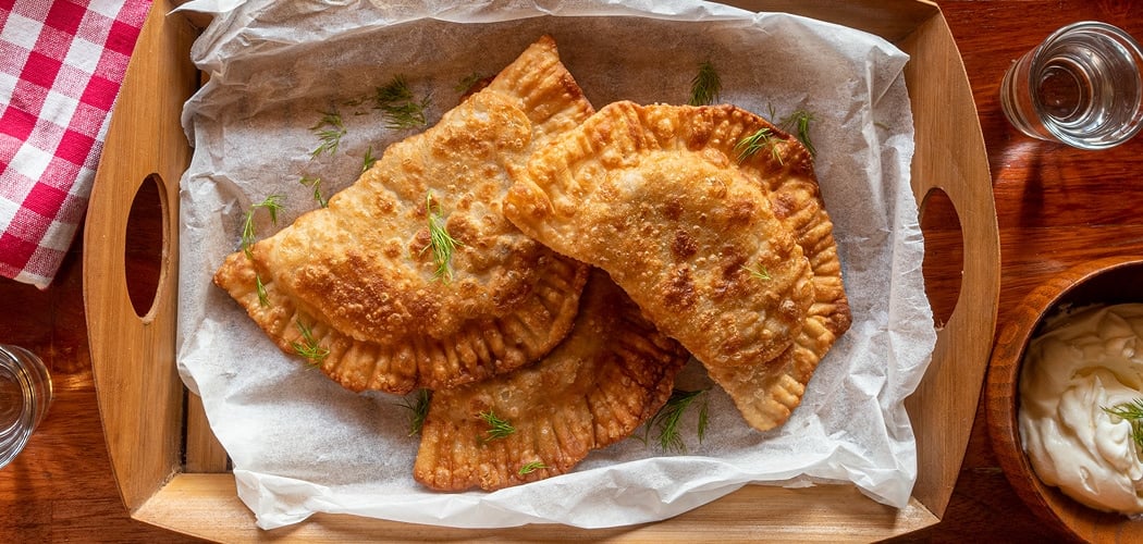 A wooden tray with fried cheberekki and dill, served alongside a bowl of cream