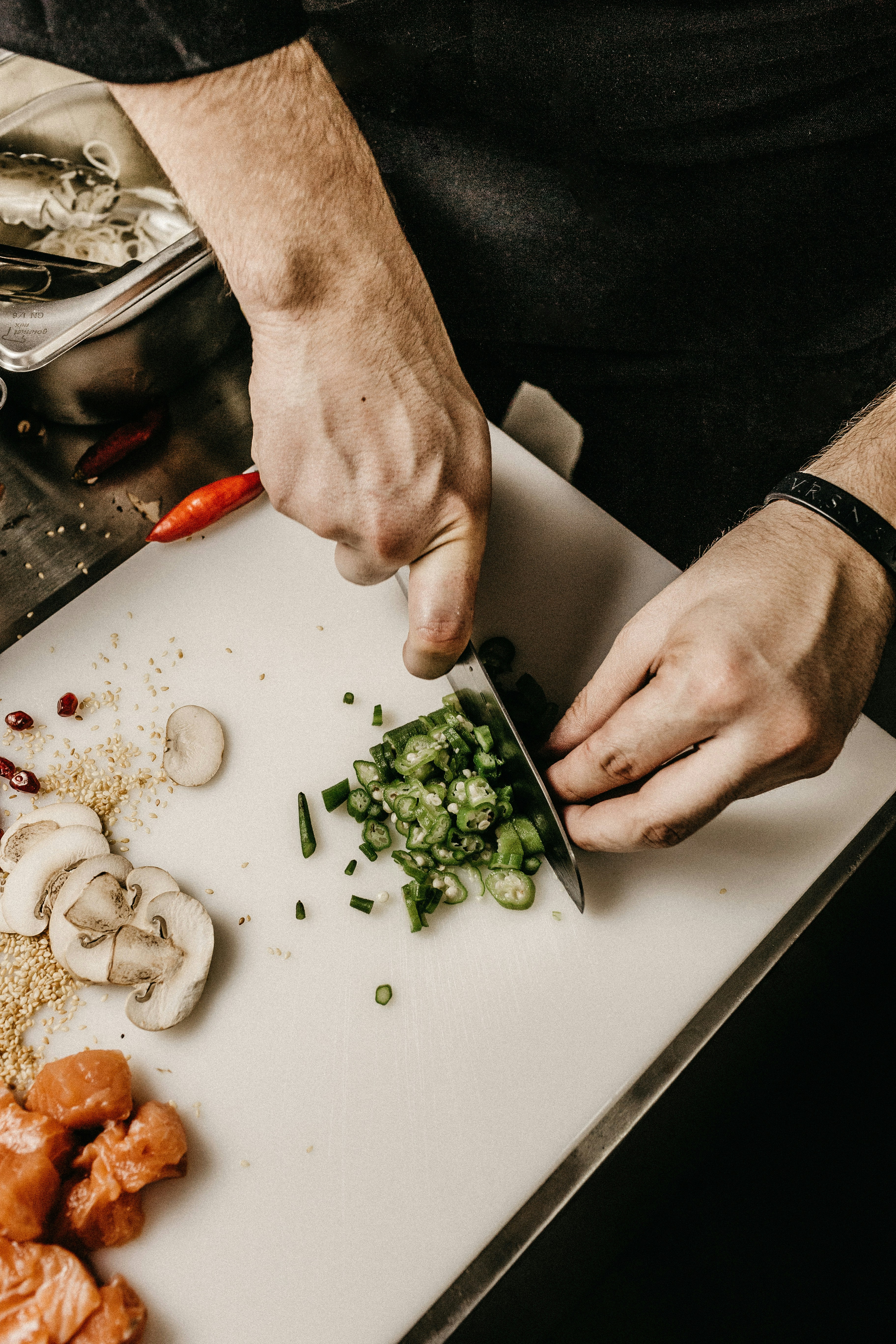 A chef dices green chilies on a white cutting board alongside sliced mushrooms and cubed meat