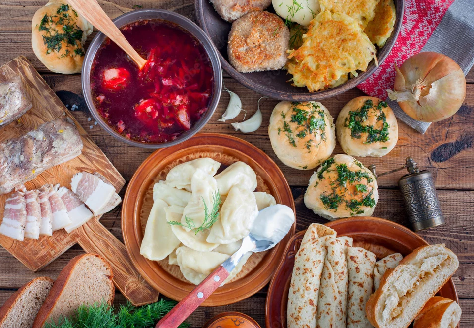 Traditional Ukrainian feast featuring borscht soup, varenyky dumplings, herb-topped buns, potato pancakes, and sliced cured meats arranged on table.