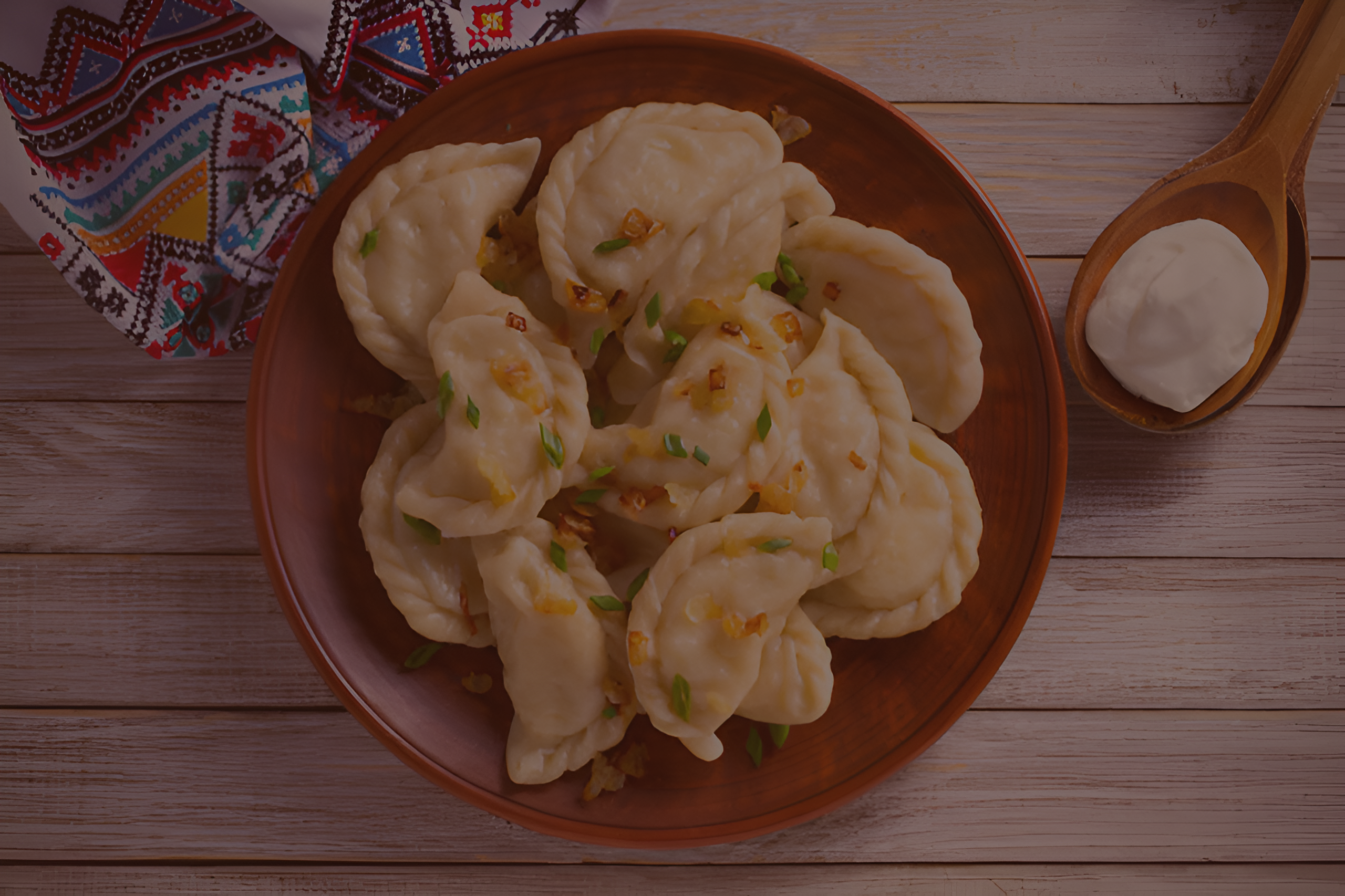 A wooden bowl of dumplings garnished with green onions sits on a table alongside a wooden spoon of sour cream and a colorful folk-patterned cloth